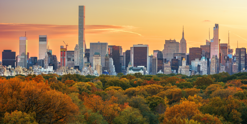 Manhattan skyline at sunset seen across Central Park in fall, with a canopy of orange, gold, and green trees in the foreground. Midtown towers, including the slender 432 Park Avenue and the Empire State Building, rise against a sky fading from purple to golden orange, with the green-roofed Plaza Hotel visible along the park's edge.