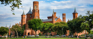 The Smithsonian Castle on the National Mall in Washington, DC, framed by lush green trees beneath a dramatic blue sky with scattered clouds.