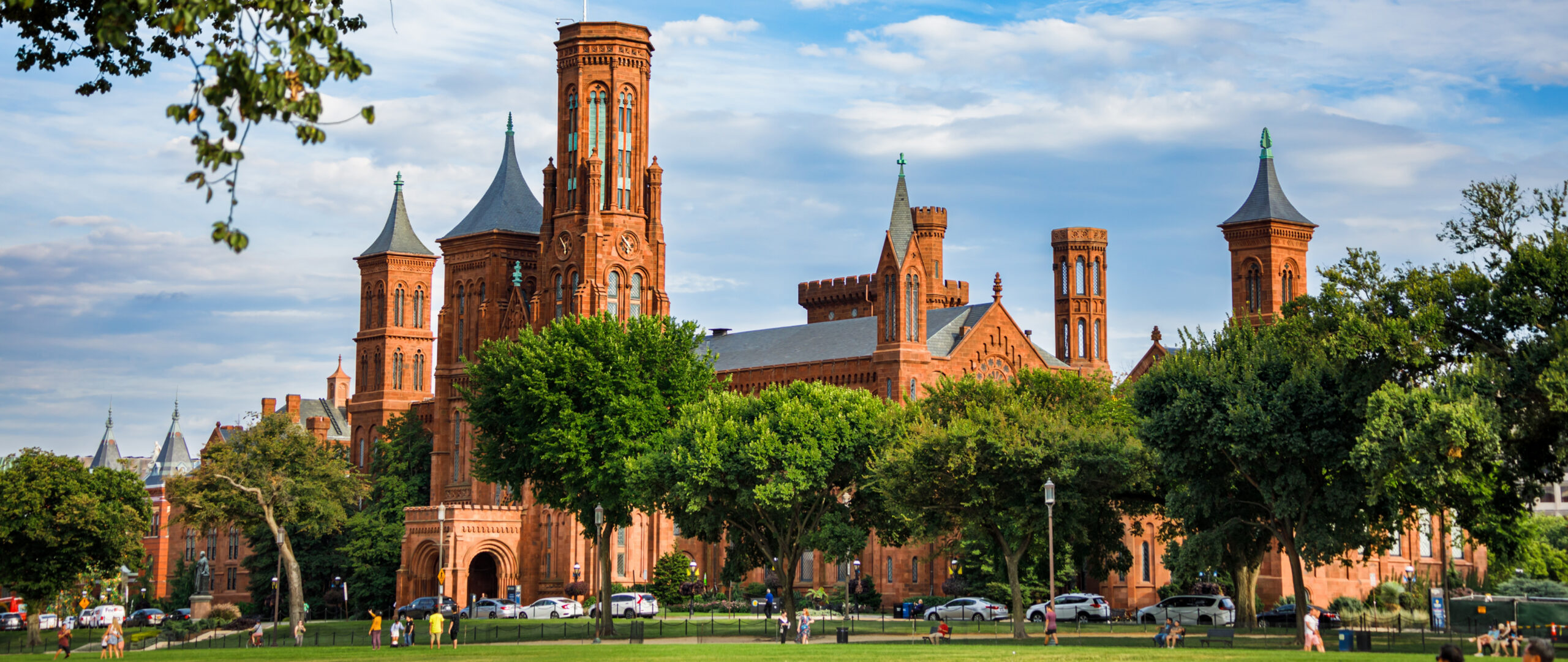 The Smithsonian Castle on the National Mall in Washington, DC, framed by lush green trees beneath a dramatic blue sky with scattered clouds.