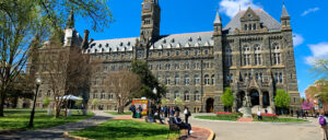 Wide view of Healy Hall at Georgetown University in Washington, DC, featuring its historic Gothic Revival stone façade, clock tower, and spires. Students and visitors walk across the front lawn and pathways on a bright, sunny day beneath a clear blue sky.