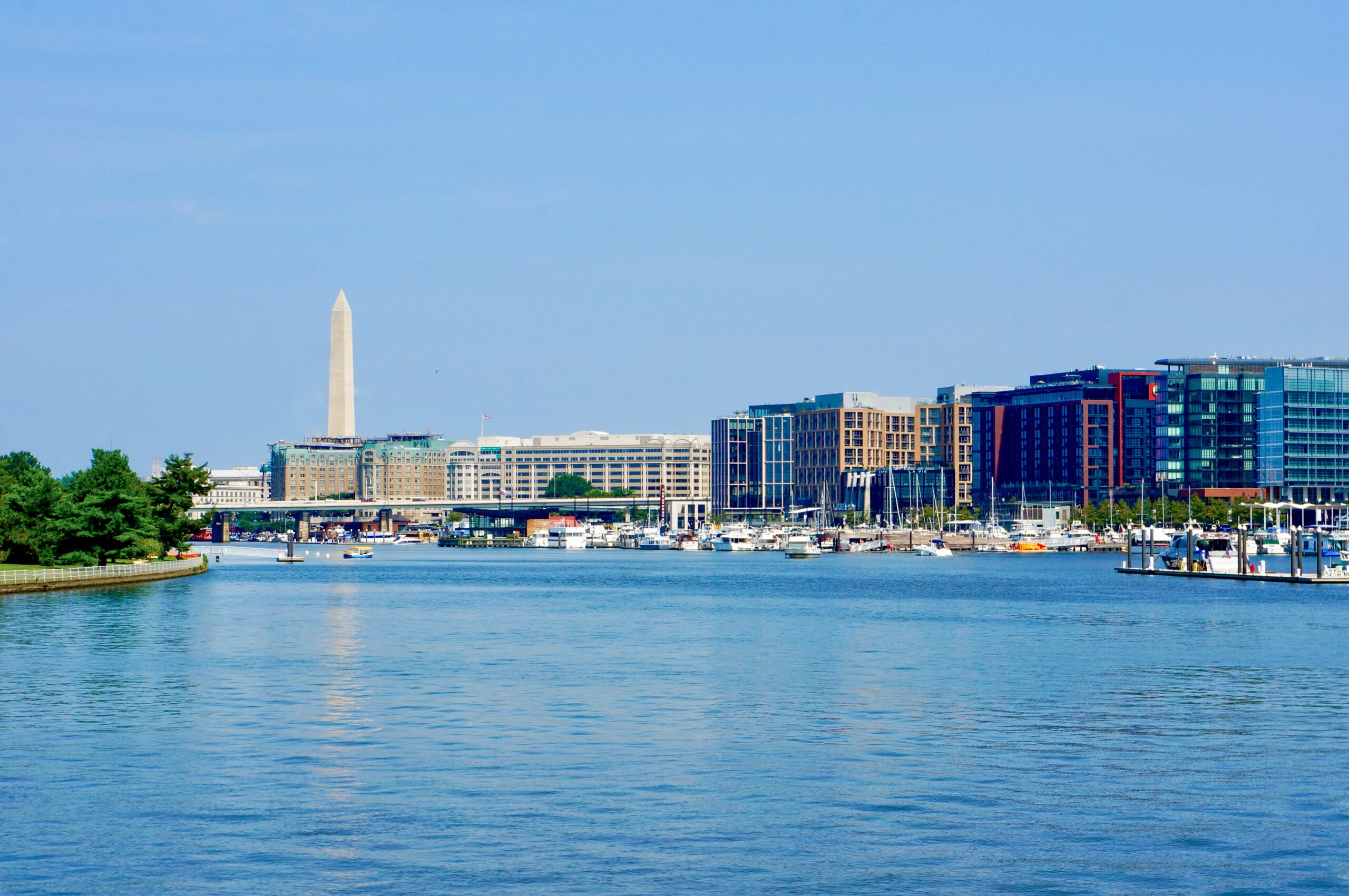 A waterfront view of The Wharf in Washington, DC, with boats docked along the marina and the Washington Monument rising in the distance.