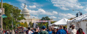 Crowds stroll along a closed-off Pennsylvania Avenue in Washington, D.C., during the Turkish Festival, passing white vendor tents and a colorful food truck. A red-and-white "Turkish Festival" banner stretches across the street, with the U.S. Capitol dome rising behind it under a bright blue sky with scattered clouds.