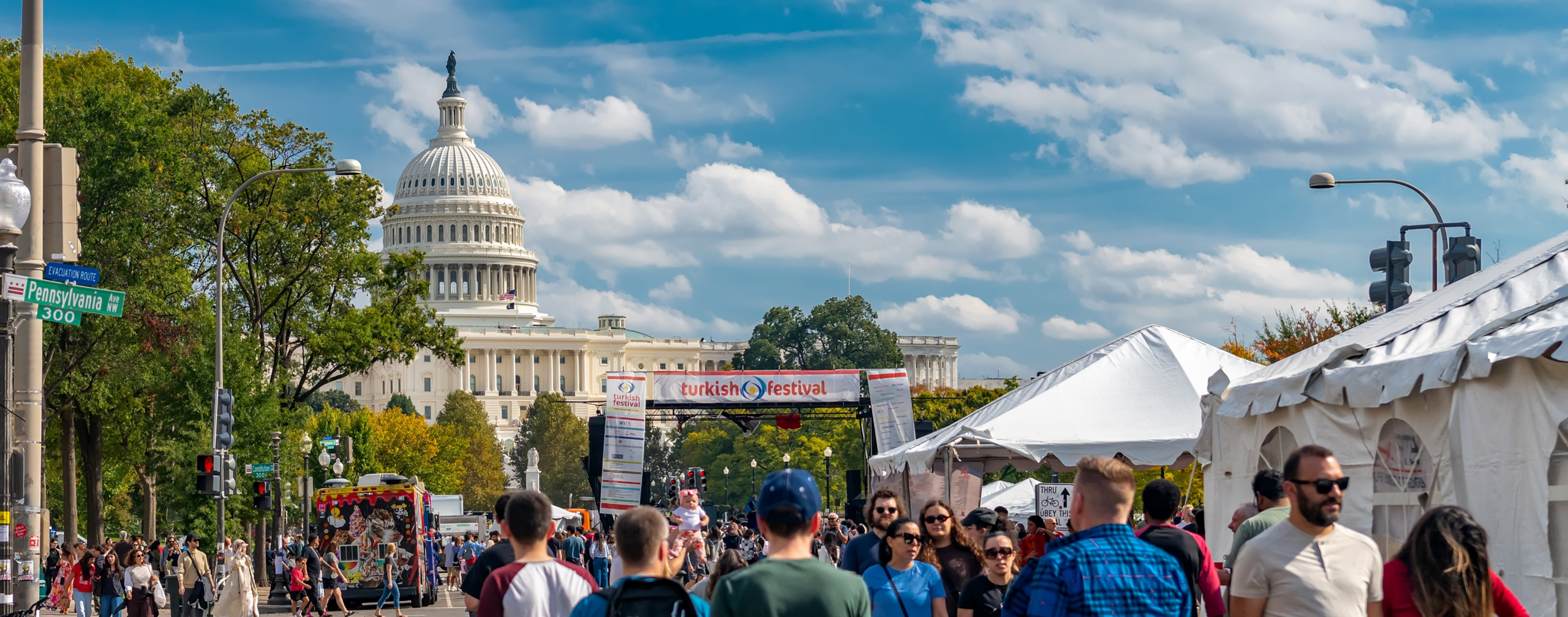 Crowds stroll along a closed-off Pennsylvania Avenue in Washington, D.C., during the Turkish Festival, passing white vendor tents and a colorful food truck. A red-and-white "Turkish Festival" banner stretches across the street, with the U.S. Capitol dome rising behind it under a bright blue sky with scattered clouds.