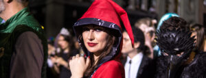 Woman in a red hooded costume with black patterned trim and red lipstick poses among costumed crowds at a Halloween night event in Washington DC, including a person in a black feathered bird mask.
