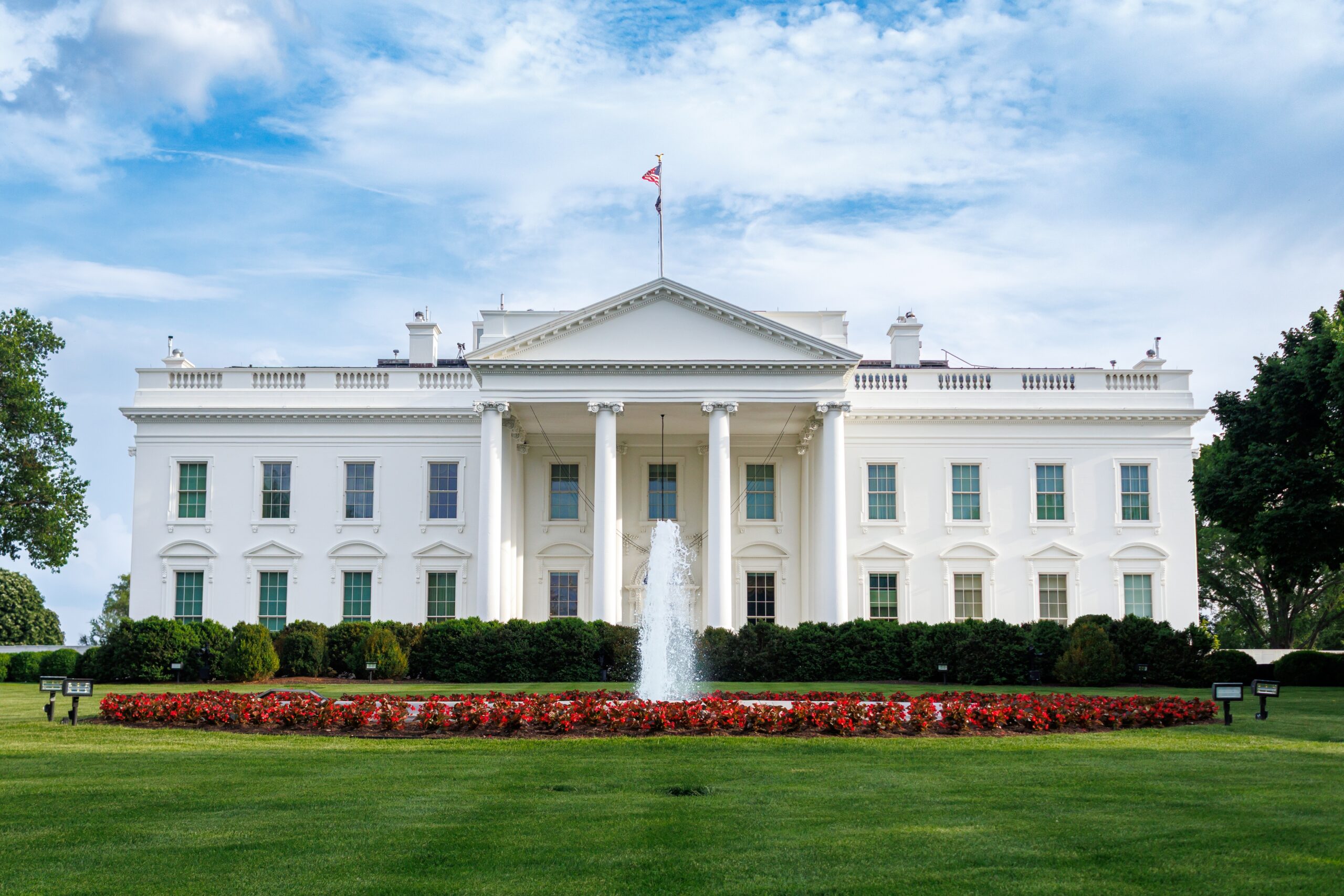 North facade of the White House in Washington, DC, with its columned North Portico, a fountain, red flower beds, and a green lawn under a blue sky.