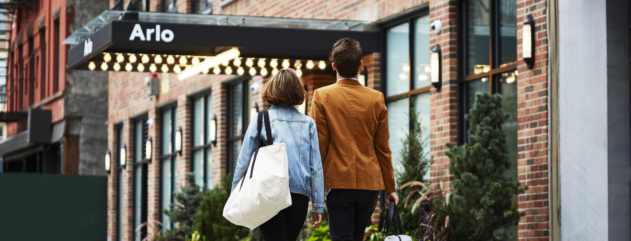 Two people walking towards a brick building with a sign reading "Arlo." The person on the left has a white bag and denim jacket, and the person on the right wears a brown jacket.