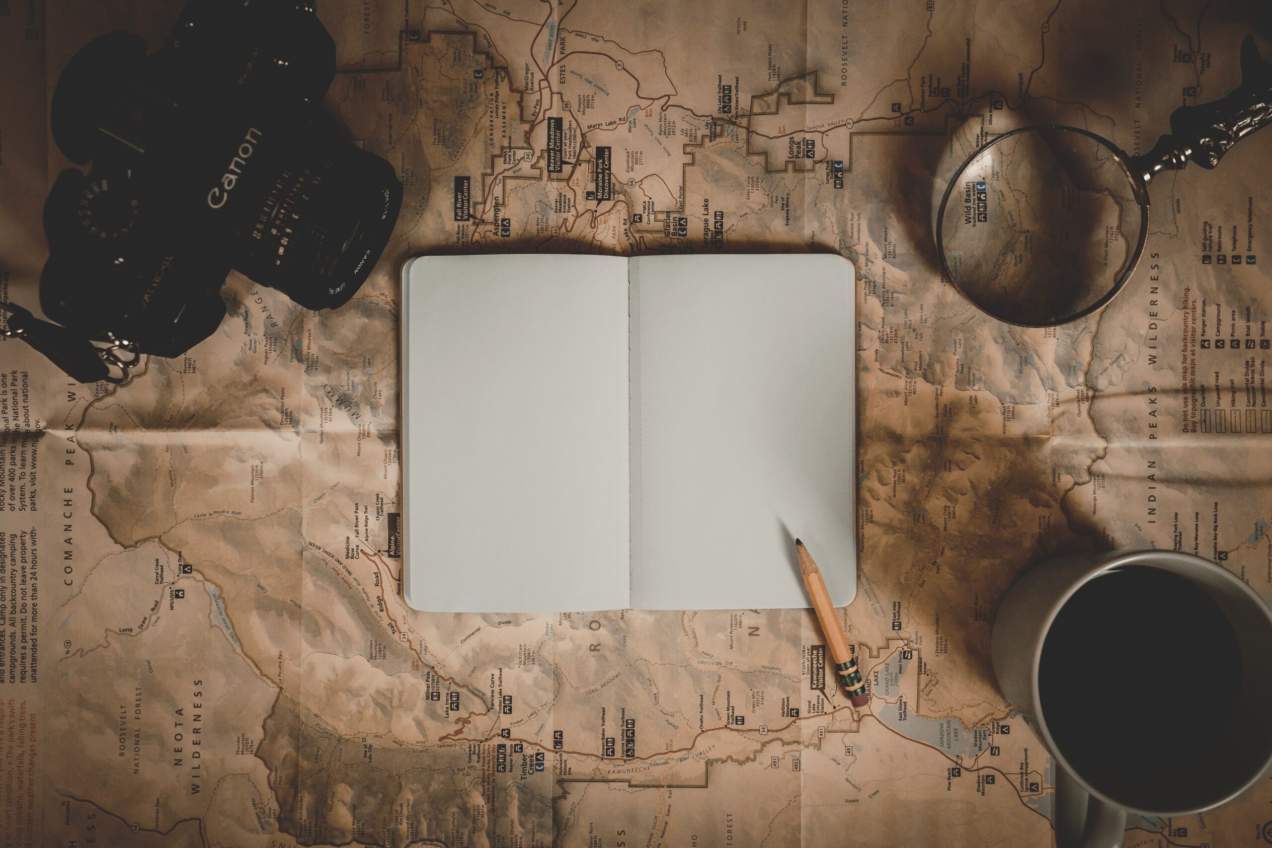 An open blank notebook, a pencil, a camera, a magnifying glass, and a coffee cup are placed on a detailed map.