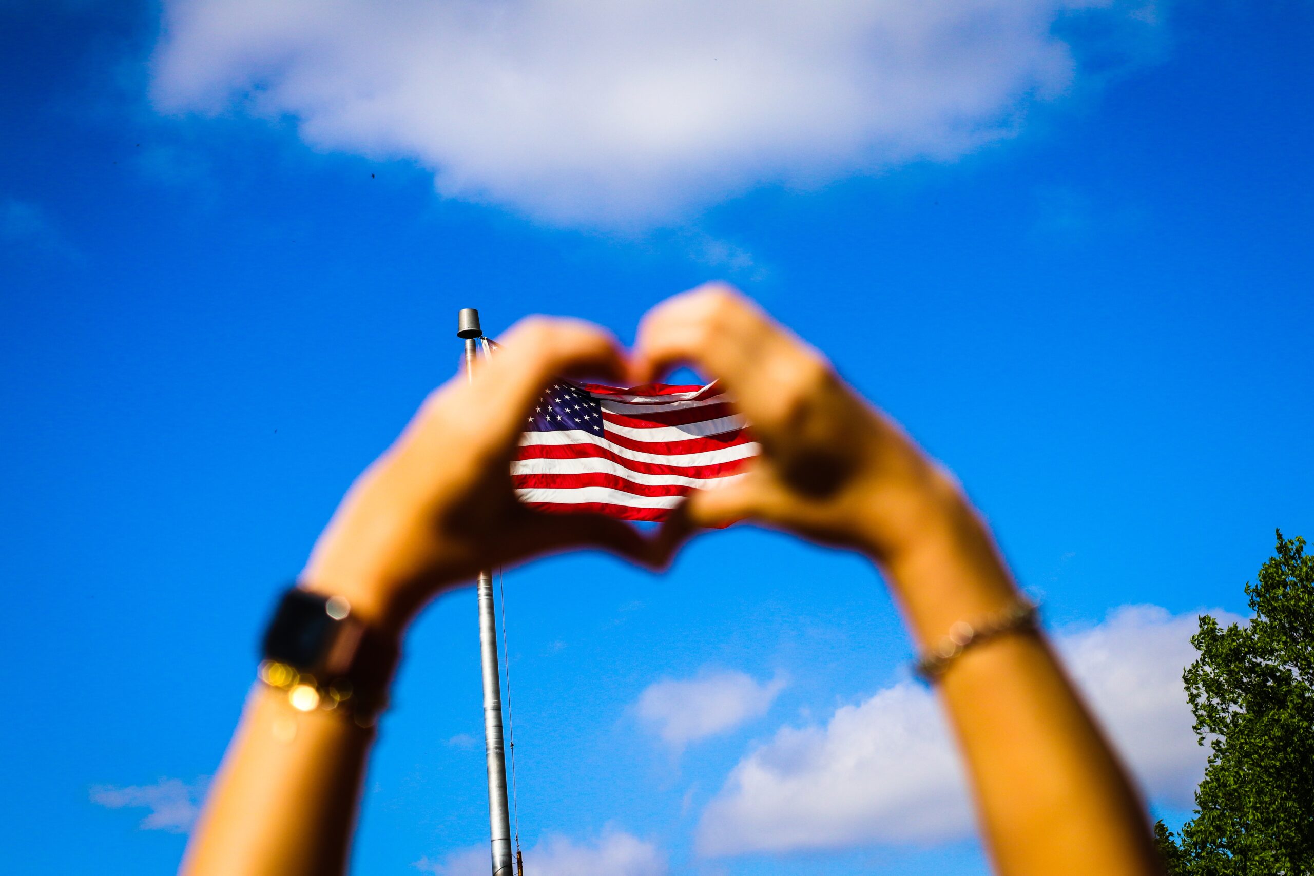 Hands forming a heart shape with a U.S. flag in the background against a clear blue sky.