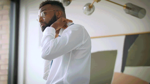 A man in a white shirt and glasses is adjusting his tie in a well-lit room with modern decor.