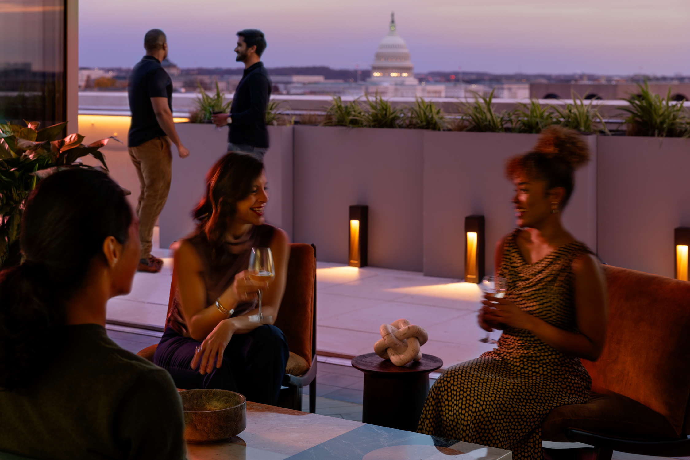 People enjoying drinks on a rooftop lounge at sunset. The US Capitol is visible in the background.