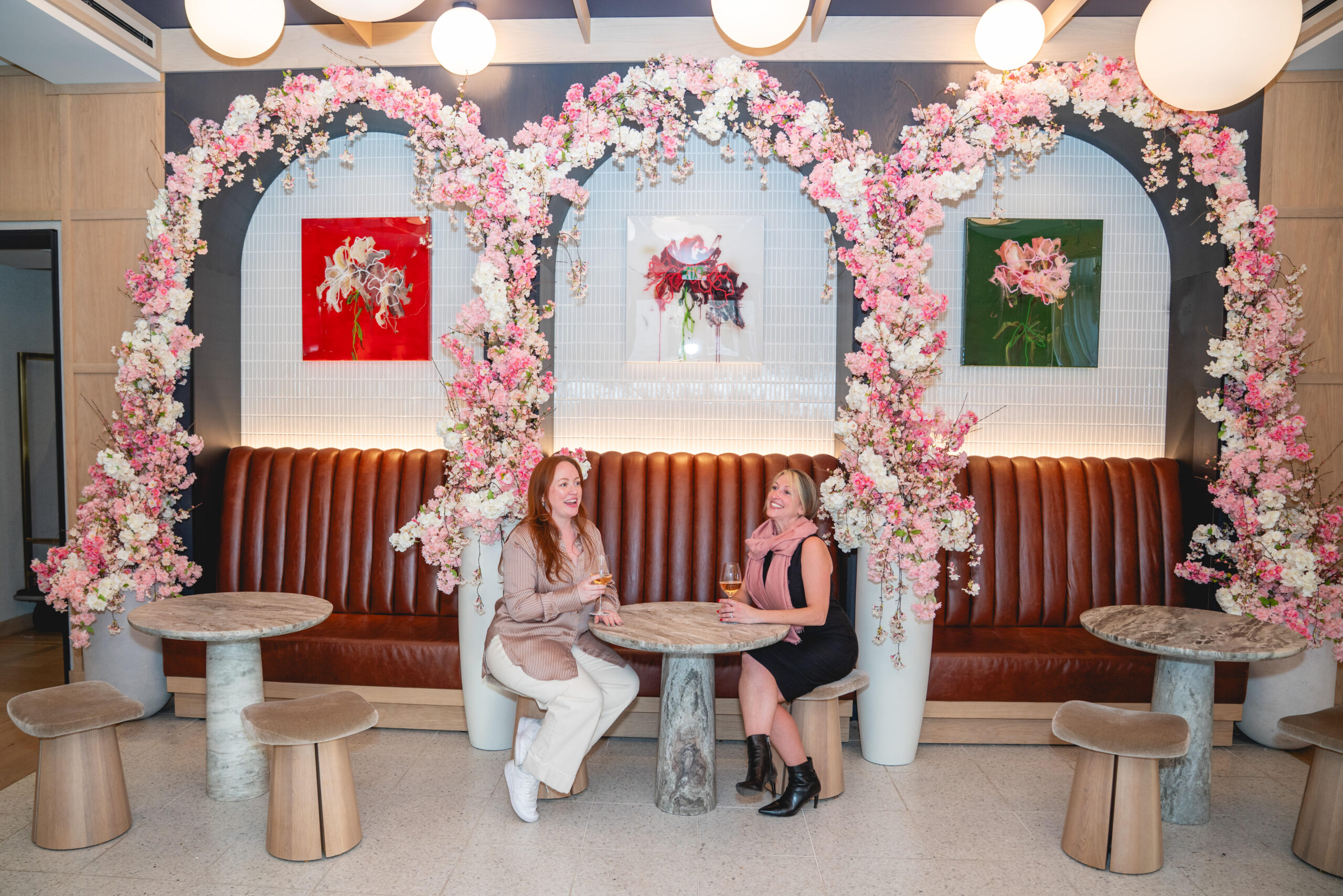 Two women sit at a round table in a café with brown booths and floral arches. Three colorful flower art pieces hang on the wall behind them.
