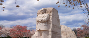 The Martin Luther King, Jr. Memorial carved into stone stands tall under a blue sky, framed by blooming cherry blossom trees in the foreground and background.