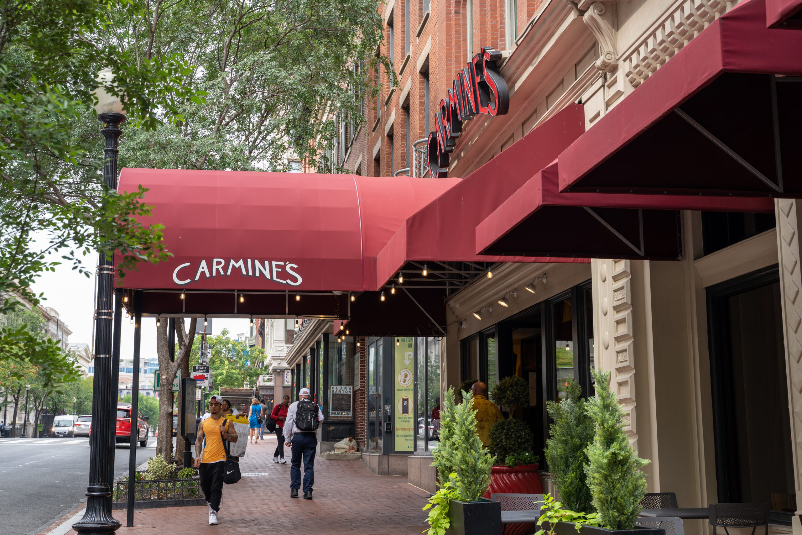 Exterior of Carmine’s Italian Restaurant in D.C. featuring signature red awnings over a brick sidewalk lined with green trees.