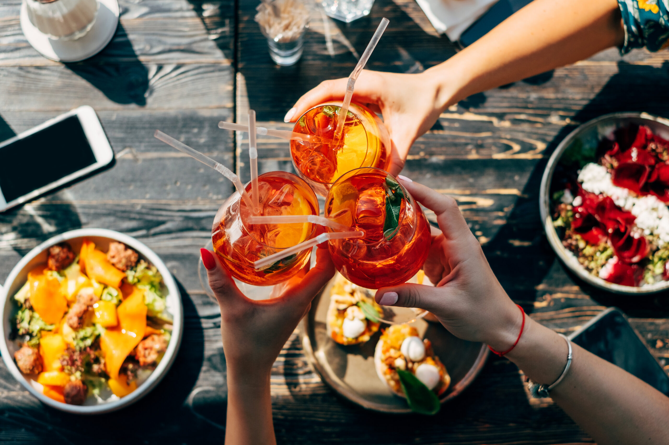 An overhead view of three people clinking bright orange cocktails over a wooden table at one of the many DC restaurants featuring outdoor dining.