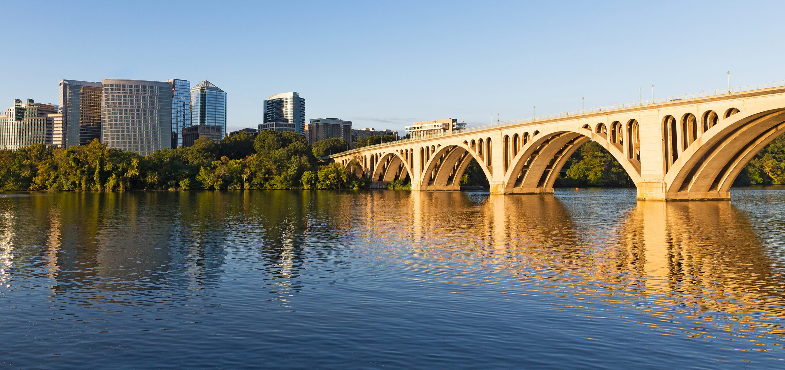A wide-angle, golden hour view of the Key Bridge spanning the Potomac River, connecting Georgetown to the Rosslyn skyline in Arlington, Virginia. The concrete arch bridge stretches across the right side of the frame, its reflection shimmering on the calm blue water. On the left bank, dense green trees line the waterfront, with modern glass skyscrapers rising behind them under a clear, pale blue sky.