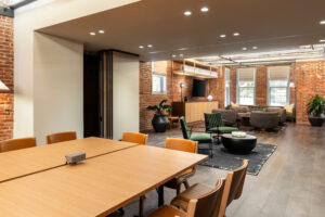 Spacious studio lounge at Arlo Washington DC featuring a large communal worktable with built-in power outlets, comfortable seating areas, exposed brick walls, modern lighting, lush plants, and a contemporary living room with a TV, creating a stylish space for working, relaxing, or socializing.
