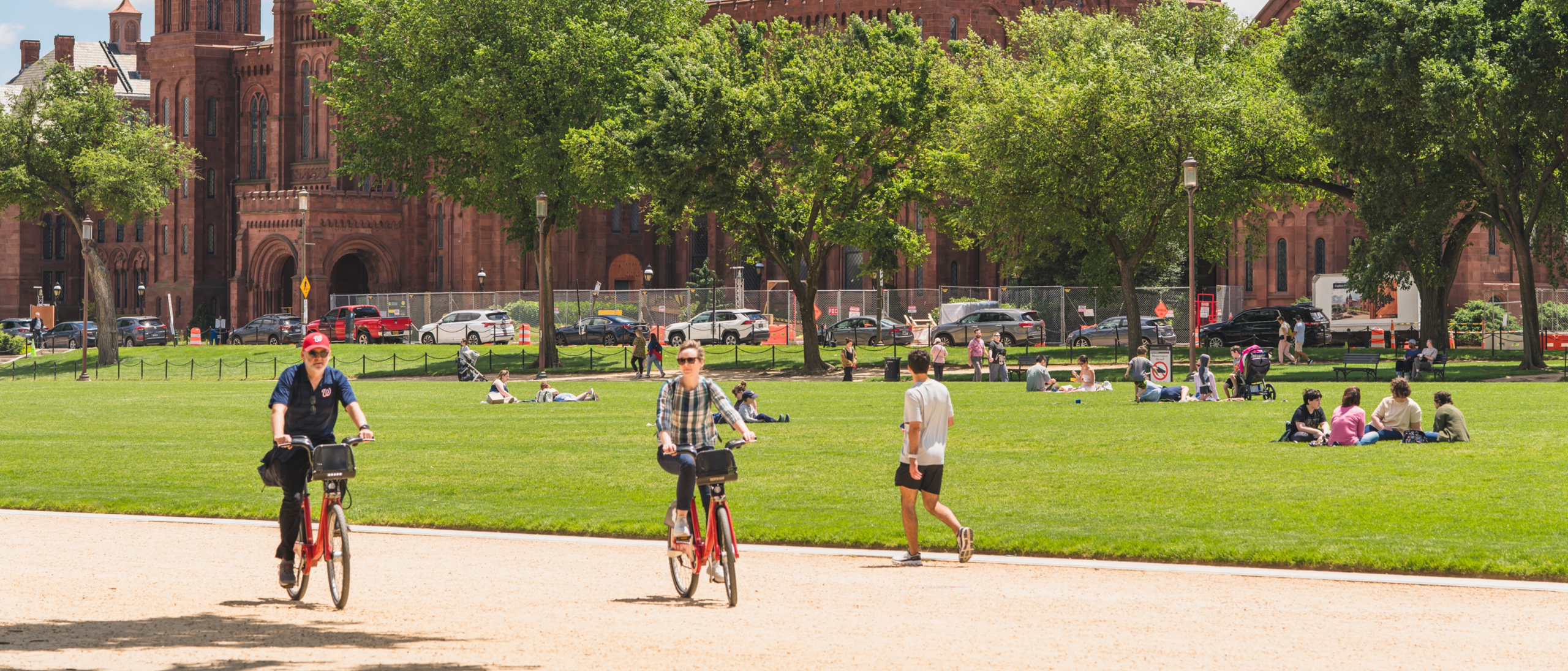 Visitors relax on the National Mall while cyclists ride past the Smithsonian Castle, the historic red sandstone building surrounded by green lawns and mature trees on a sunny day.