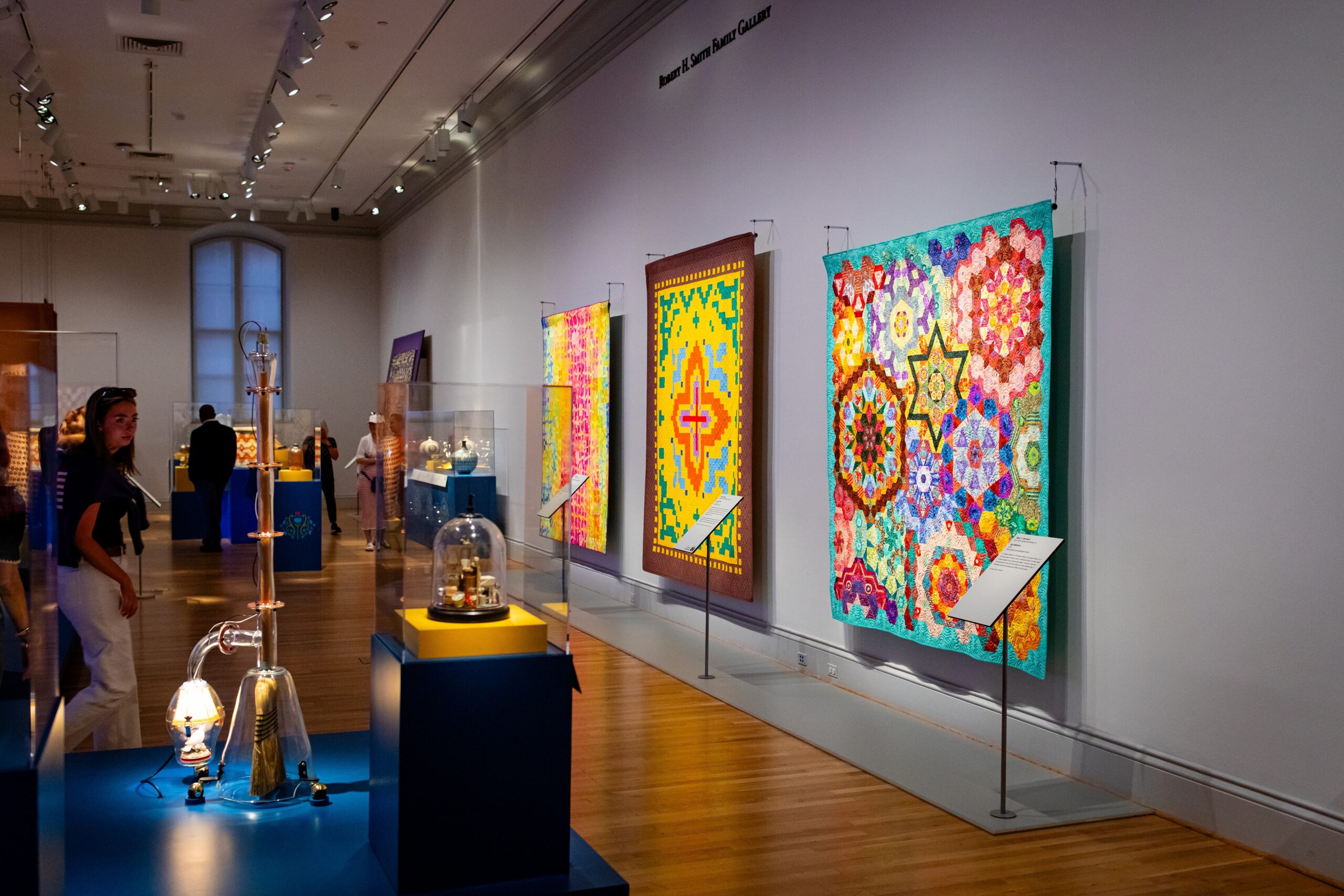 Interior of a museum gallery displaying colorful textile artworks and historical artifacts in glass cases, with visitors viewing the exhibits.