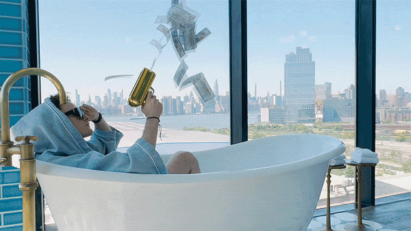 A person in a bathrobe sits in a bathtub next to large windows and uses a gold-colored device to blast cash into the air. City buildings are visible in the background.