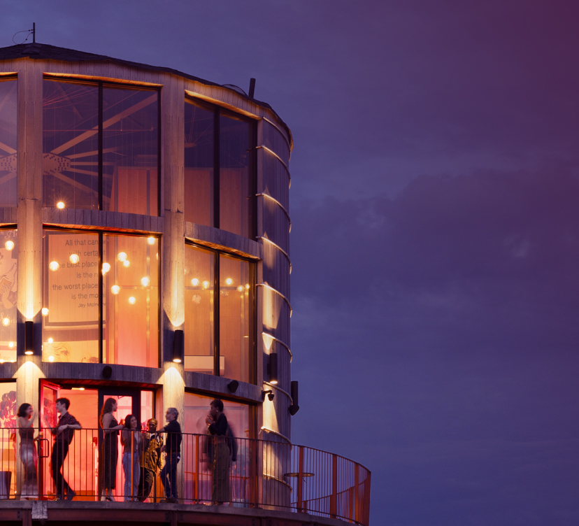 People standing inside and on the balcony of a circular glass building, illuminated by interior lights, against a dusky sky.