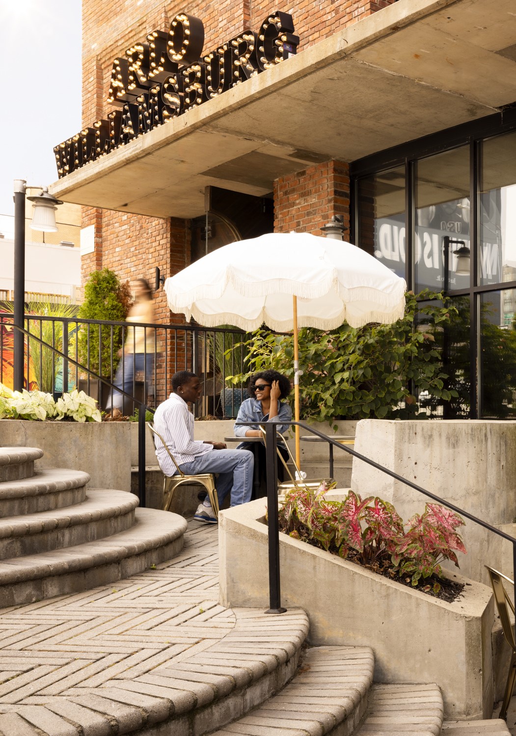 Two people sit at an outdoor table under a white umbrella by the entrance of a brick building labeled "Arlo Williamsburg," with curved steps and greenery nearby.