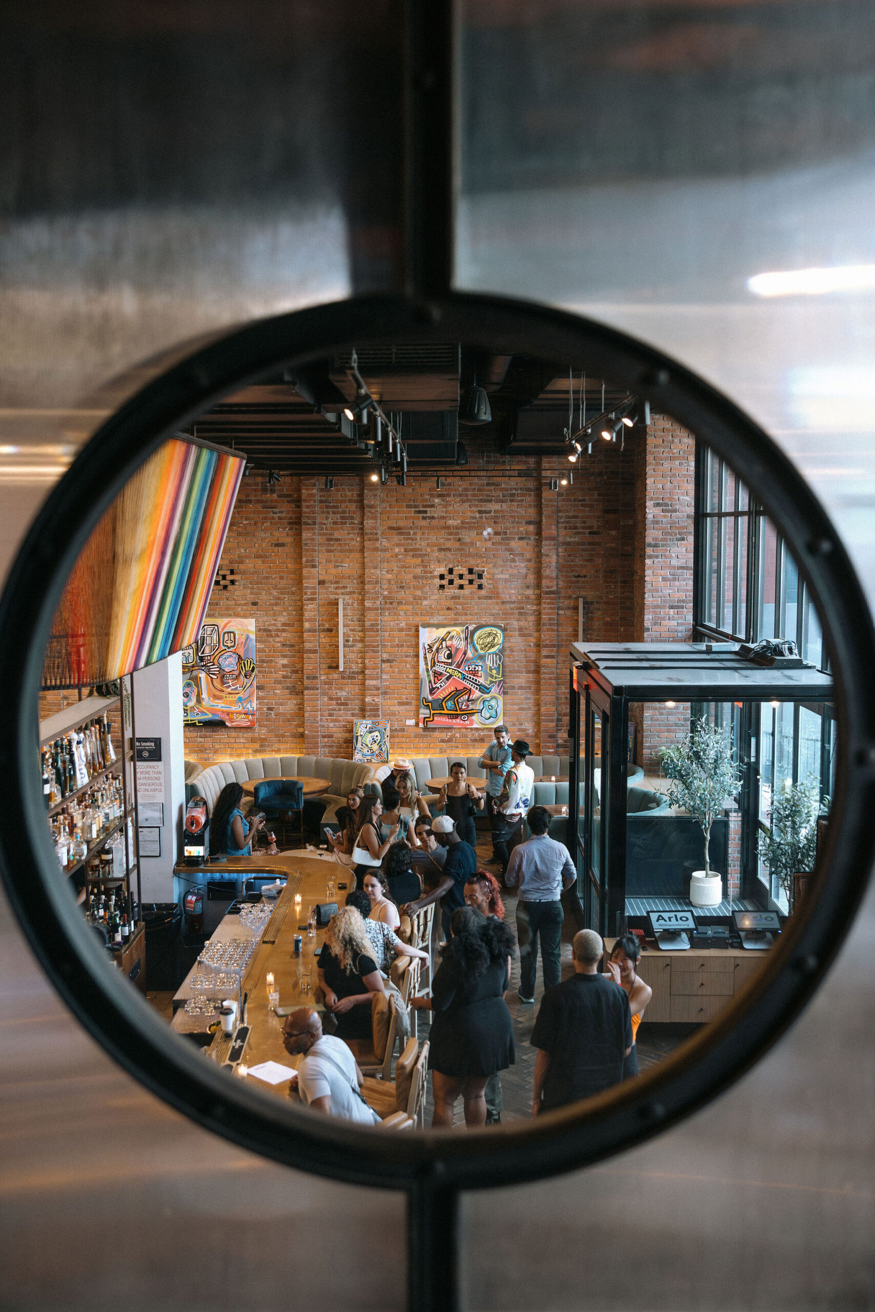 People gather and converse around a bar in a modern, brick-walled restaurant, seen through a circular window. Colorful artwork and a rainbow flag are visible on the walls.