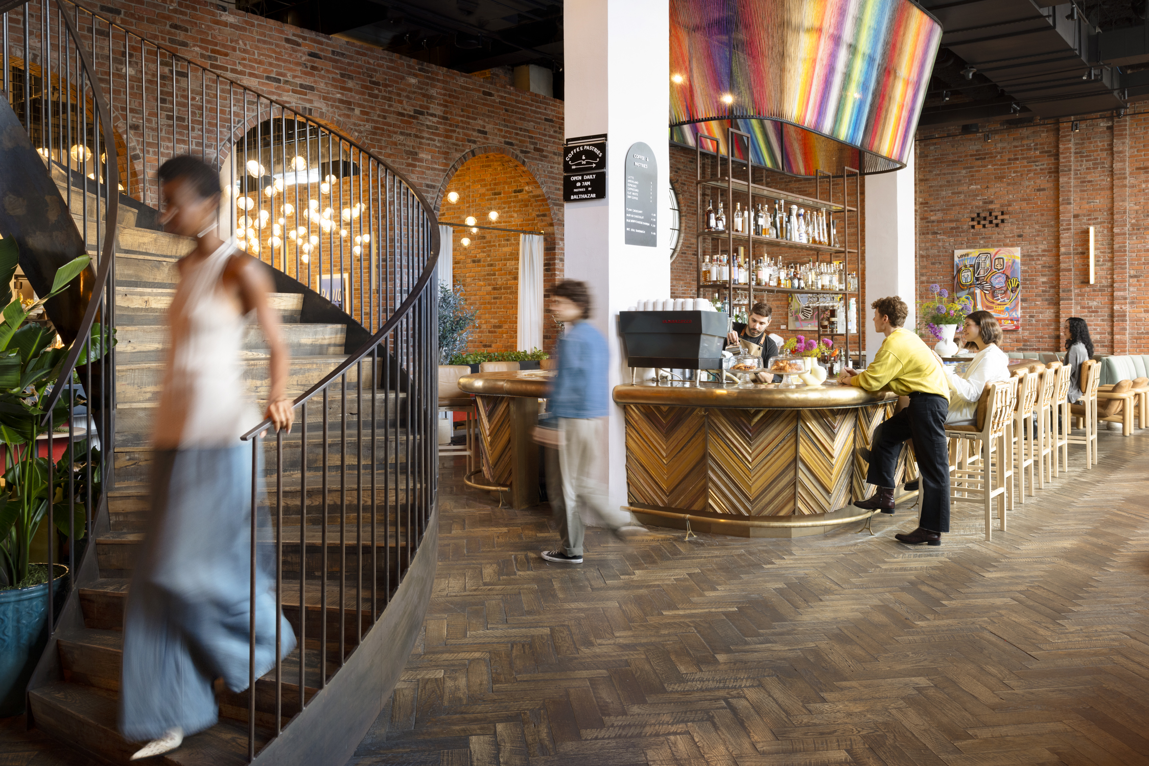 Modern café with a curved staircase, people sitting at the bar, and others walking; brick walls, wood flooring, and colorful decor visible.