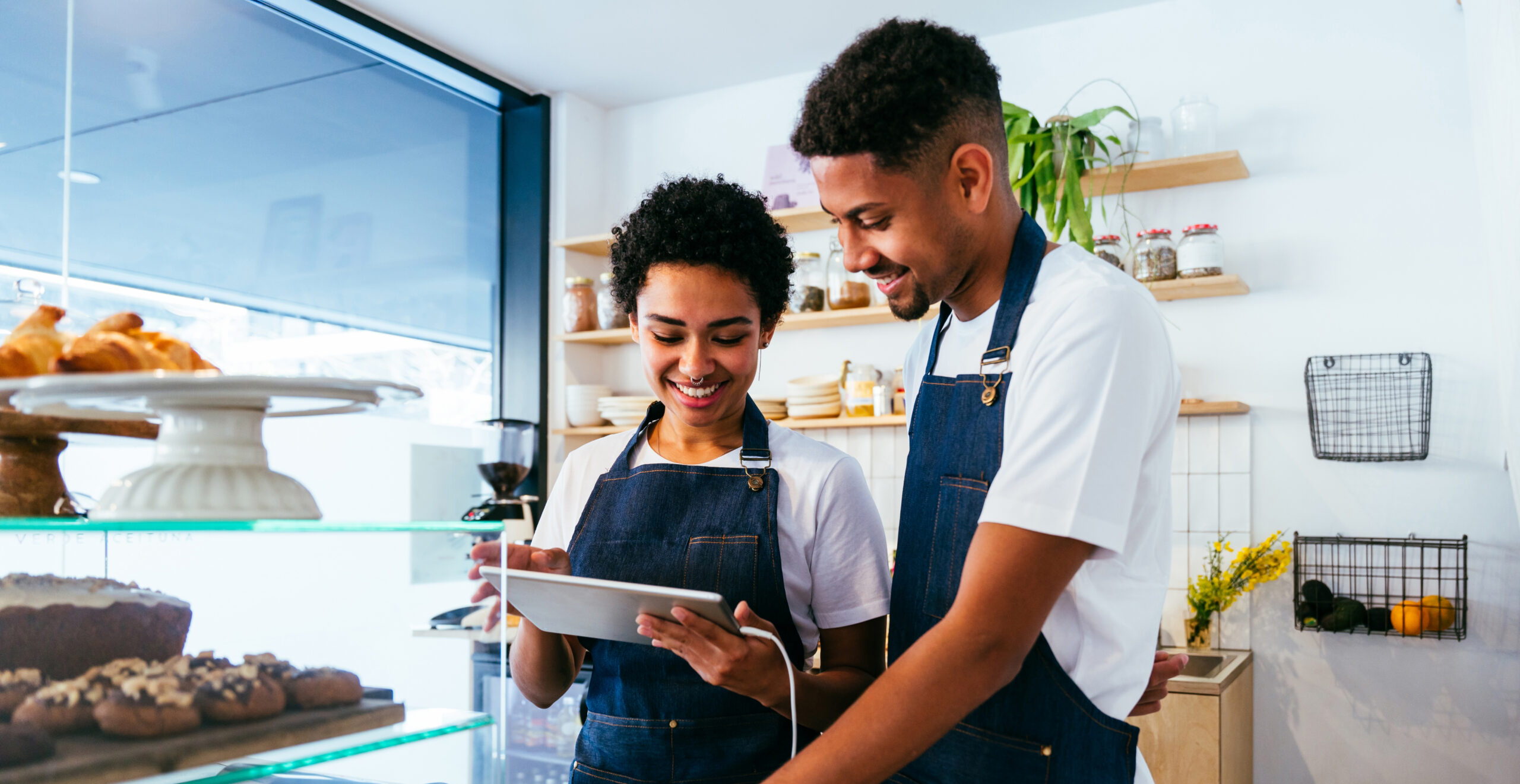 Two young Black entrepreneurs wearing denim aprons smile as they look at a white tablet behind the counter of a bright, modern bakery.