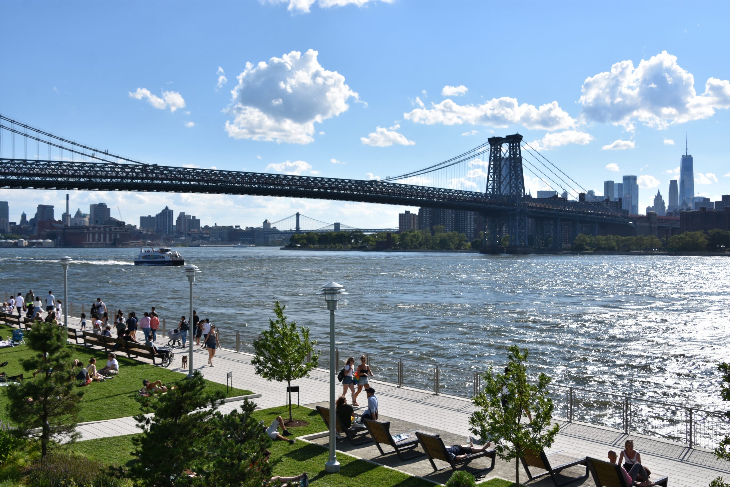 Williamsburg waterfront park with the Williamsburg Bridge, East River, and Lower Manhattan skyline on a sunny summer afternoon in Brooklyn, NYC.