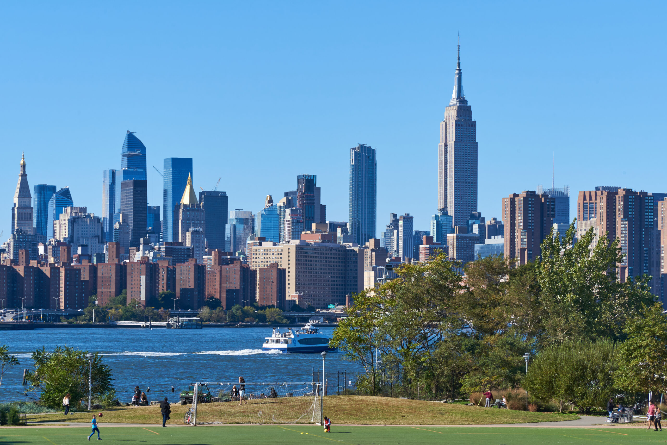 **Alt text:** View of East River State Park with a grassy lawn overlooking the East River, a ferry passing on the water, and the Manhattan skyline featuring the Empire State Building under a clear blue sky.