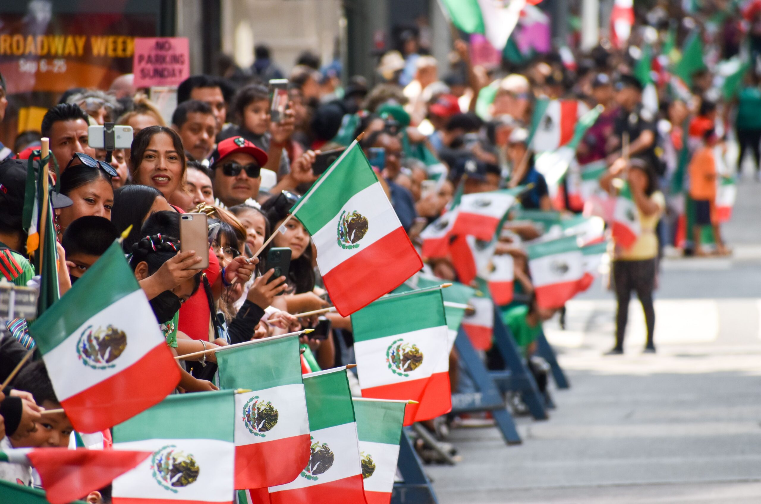 Crowds line the street waving Mexican flags during a Mexican Independence Day parade, celebrating with colorful displays, cheering spectators, and festive energy.