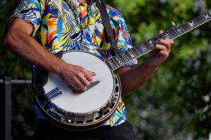 Close-up of a musician playing a banjo outdoors, wearing a colorful patterned shirt at a Brooklyn Americana music festival.