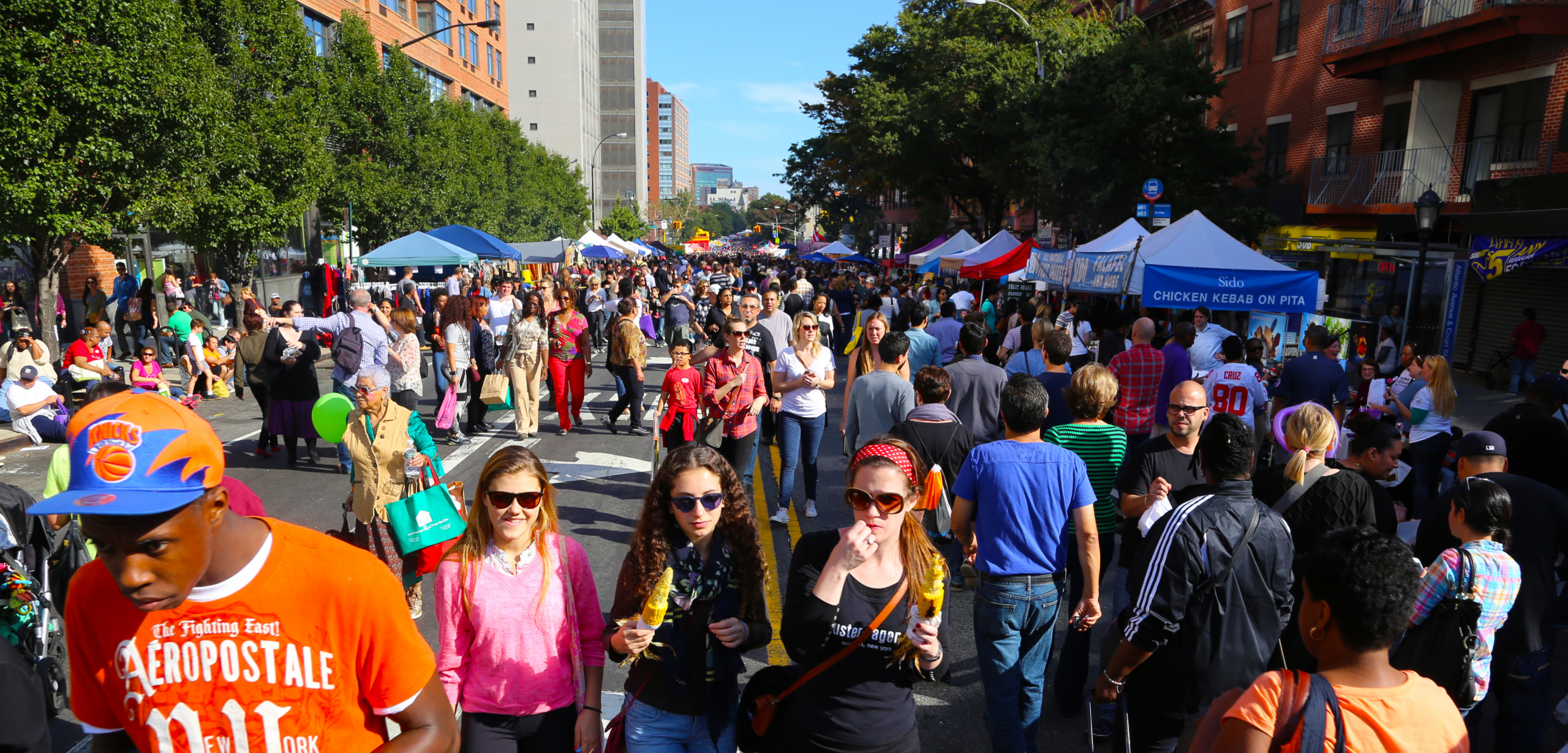 A crowded Atlantic Antic street festival in Brooklyn, with thousands of people walking between food stalls and vendor tents along the closed-off avenue.