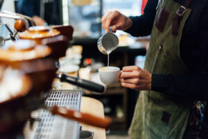 Barista in a green apron pouring steamed milk to create latte art in a white cup, next to an espresso machine.