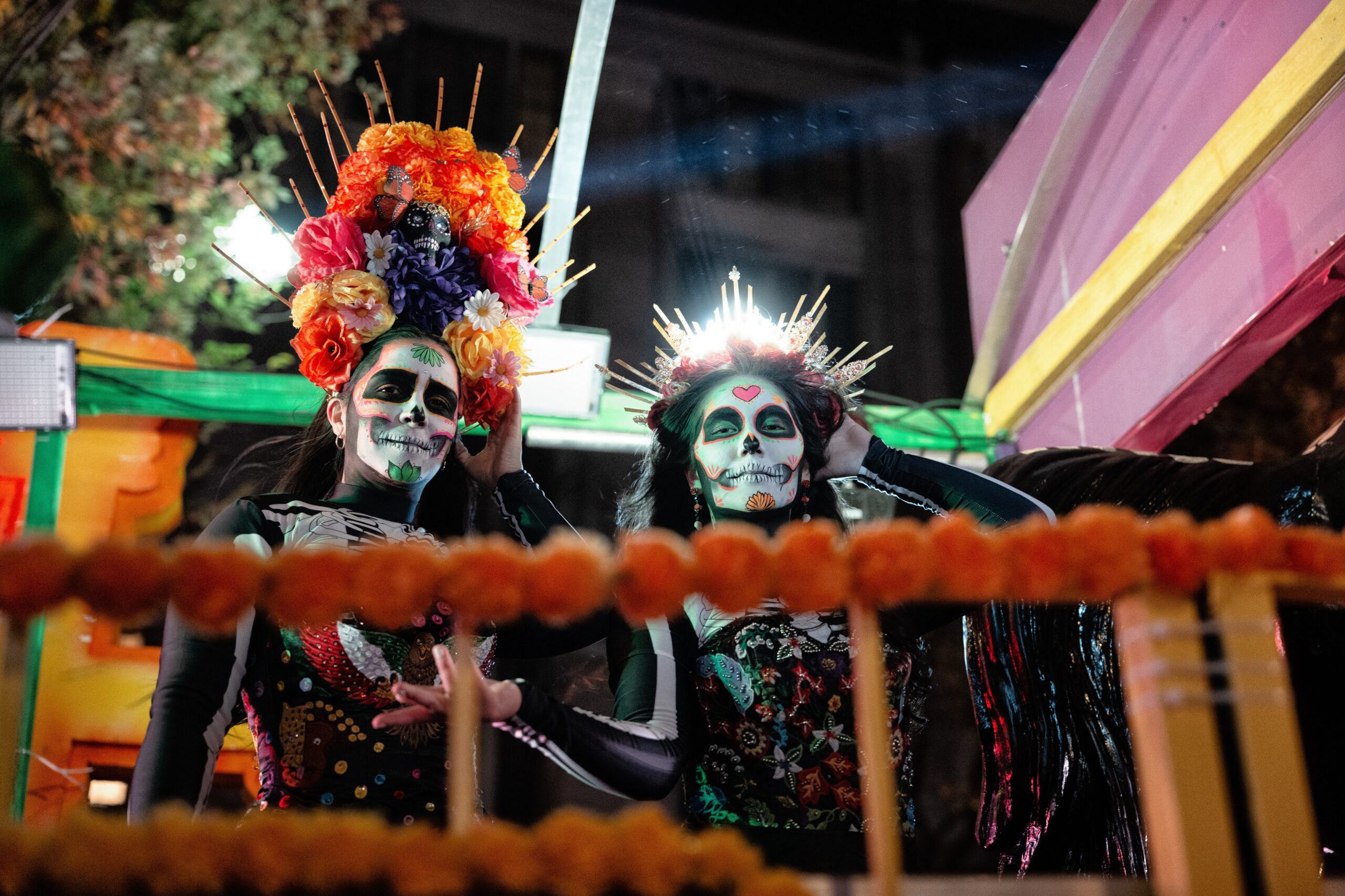 Two performers in sugar skull face paint and Day of the Dead–style costumes ride a marigold-trimmed parade float at night, one in a tall floral headdress and the other in a glowing gold crown, during Halloween in Brooklyn.
