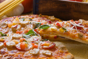 Close-up of a thin-crust pizza topped with mushrooms, pepperoni, diced peppers, fresh basil, and shredded parmesan, with a slice being lifted on a server and dry spaghetti and garlic in the background.