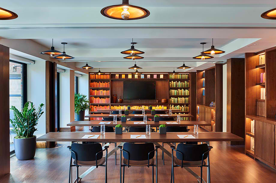 A modern conference room with long wooden tables, black chairs, bookshelves filled with colorful books, potted plants, and ceiling lights. A large screen is mounted on the wall at the front.