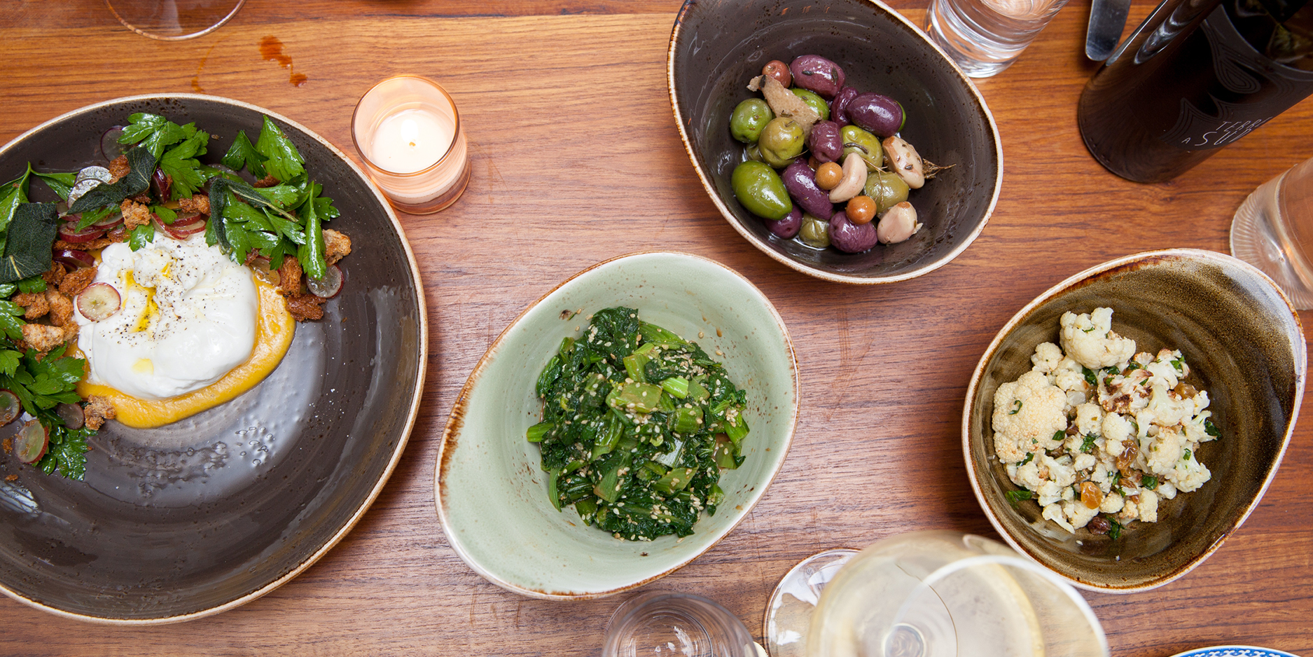 A wooden table with plates of assorted dishes including burrata with greens, a bowl of mixed olives, sautéed greens, and cauliflower. A glass of white wine and a lit candle are also present.