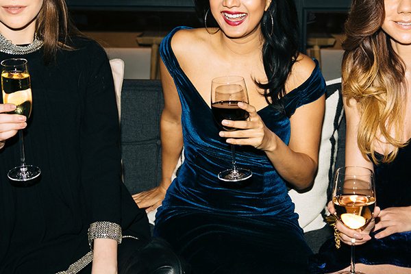 Three women sitting on a couch, wearing elegant dresses and holding wine glasses, smiling and enjoying a social event.