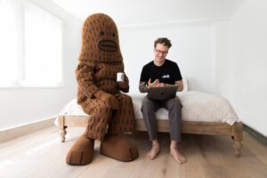 Male artist in black tshirt sitting on a bed in a room with someone dressed in a costume of a large brown stylized character similar to an ape