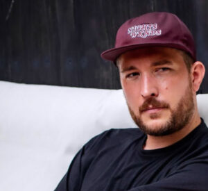 Man in black tshirt sitting down on white sofa and looking into the camera as he is wearing a maroon cap with white retro lettering that says NIGHTS WKNDS