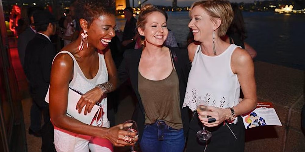 Three women, laughing at an event, holding drinks.