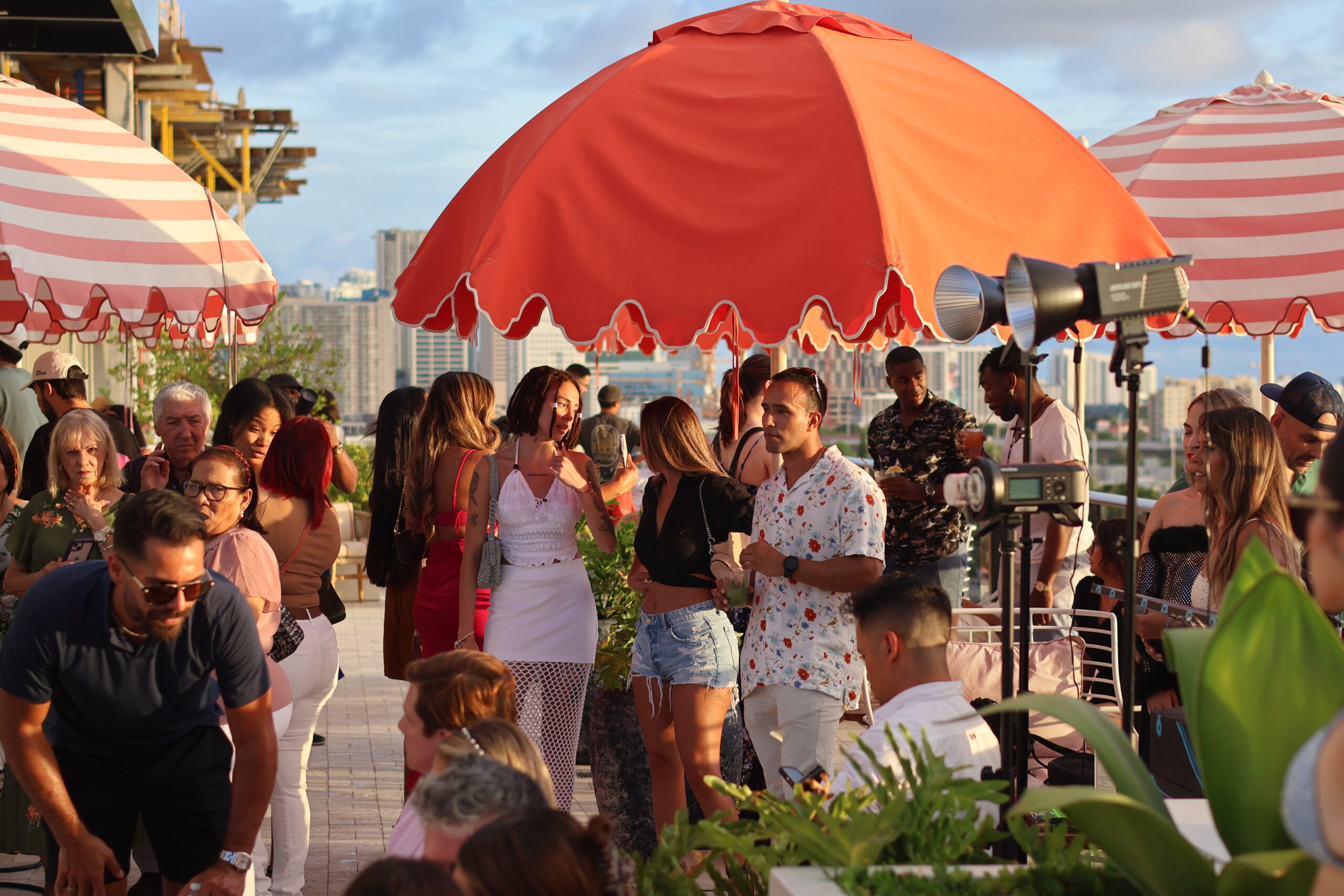 A rooftop gathering with people socializing under red and white umbrellas, set against a city skyline background.