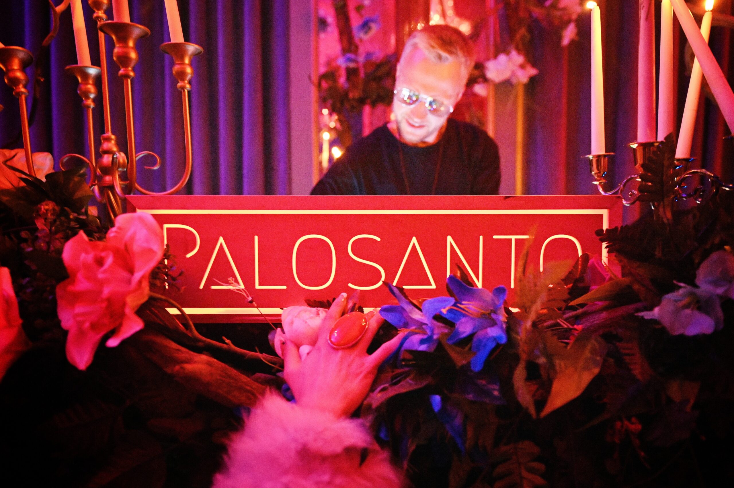 A person with reflective glasses stands behind a red sign that reads "PALO SANTO" amidst decorative flowers and candles in a dimly lit environment.