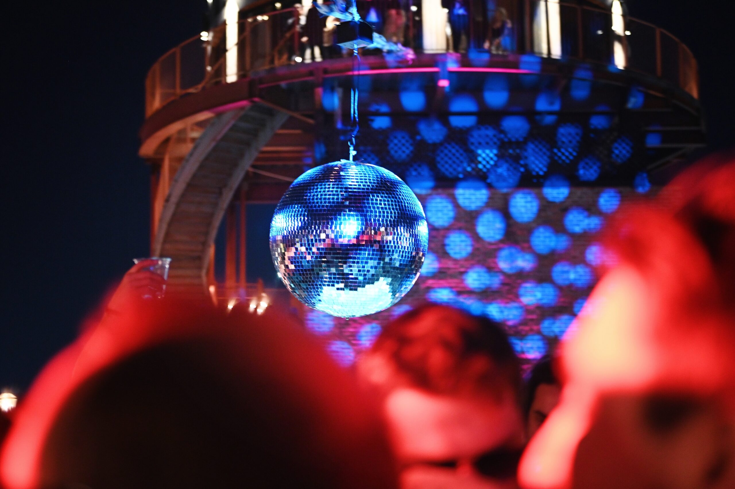 Close-up of a blue disco ball in a dimly lit room with red and blue lights, surrounded by people at a party.