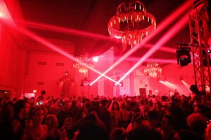 A large crowd gathers under red lighting with chandeliers and laser beams at an indoor party or nightclub event.