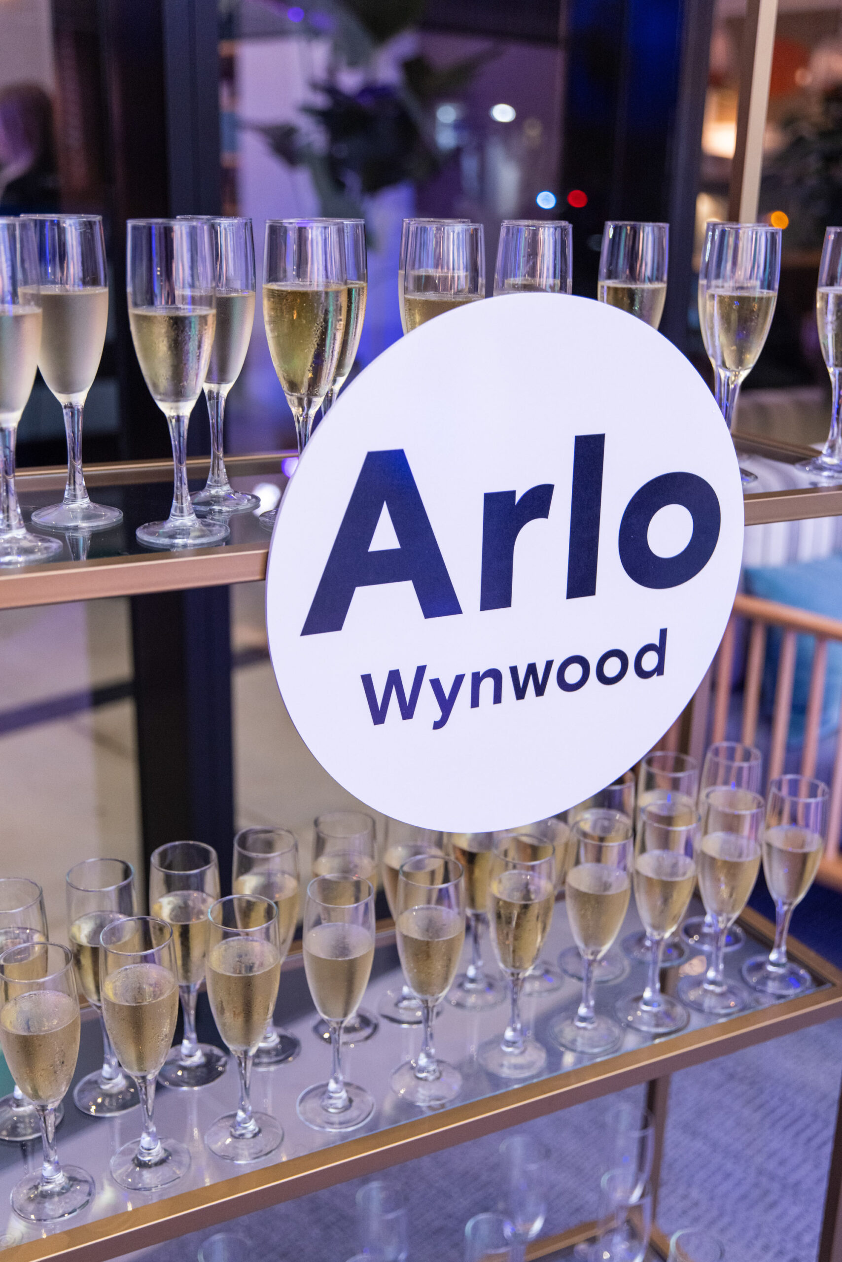 A display of champagne glasses on a bar cart at an event, with a circular sign reading "Arlo Wynwood" in the foreground.