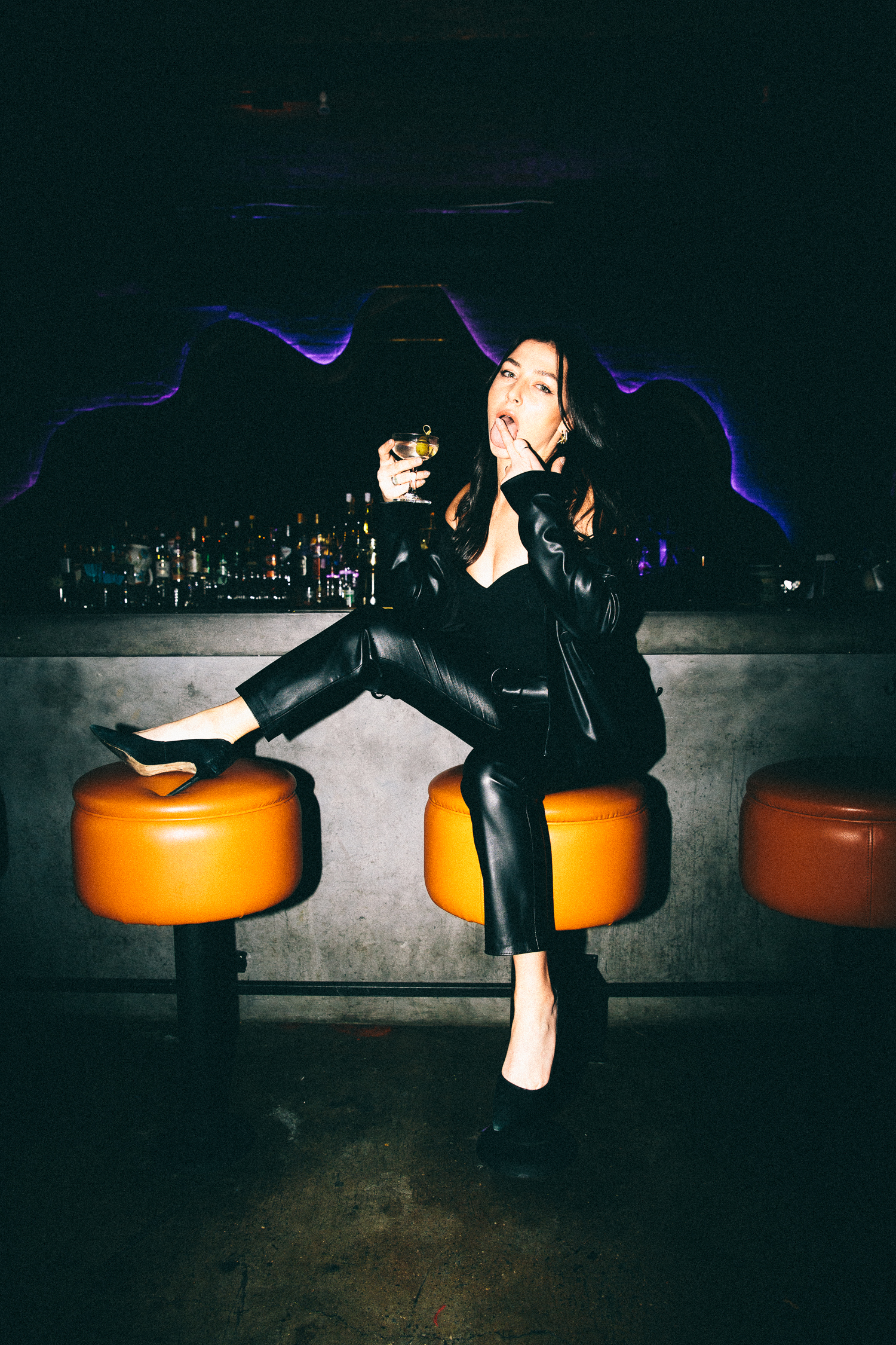 A person in black leather clothing sits on an orange barstool, holding a drink and resting one foot on another stool, with a bar and bottles visible in the background.