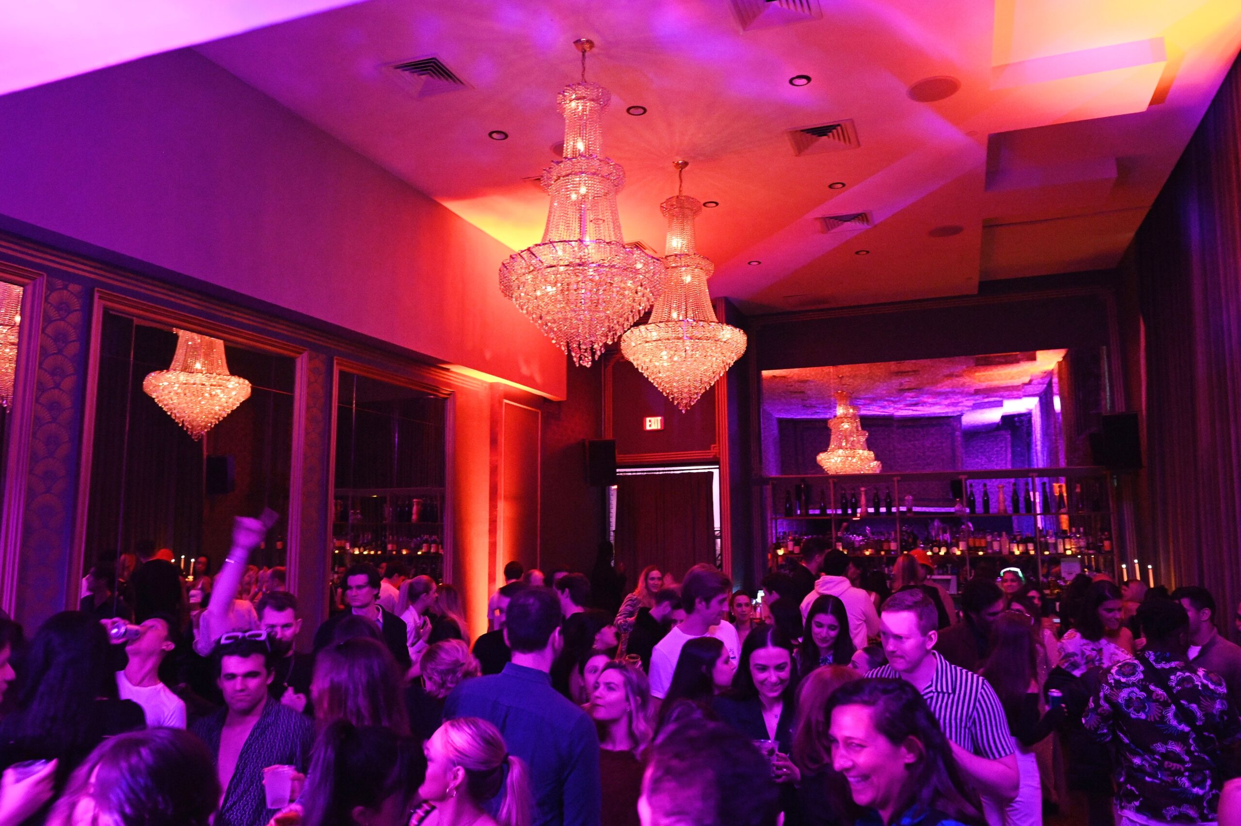 A lively crowd of people mingling and dancing in a dimly lit room with luxurious chandeliers and purple lighting.