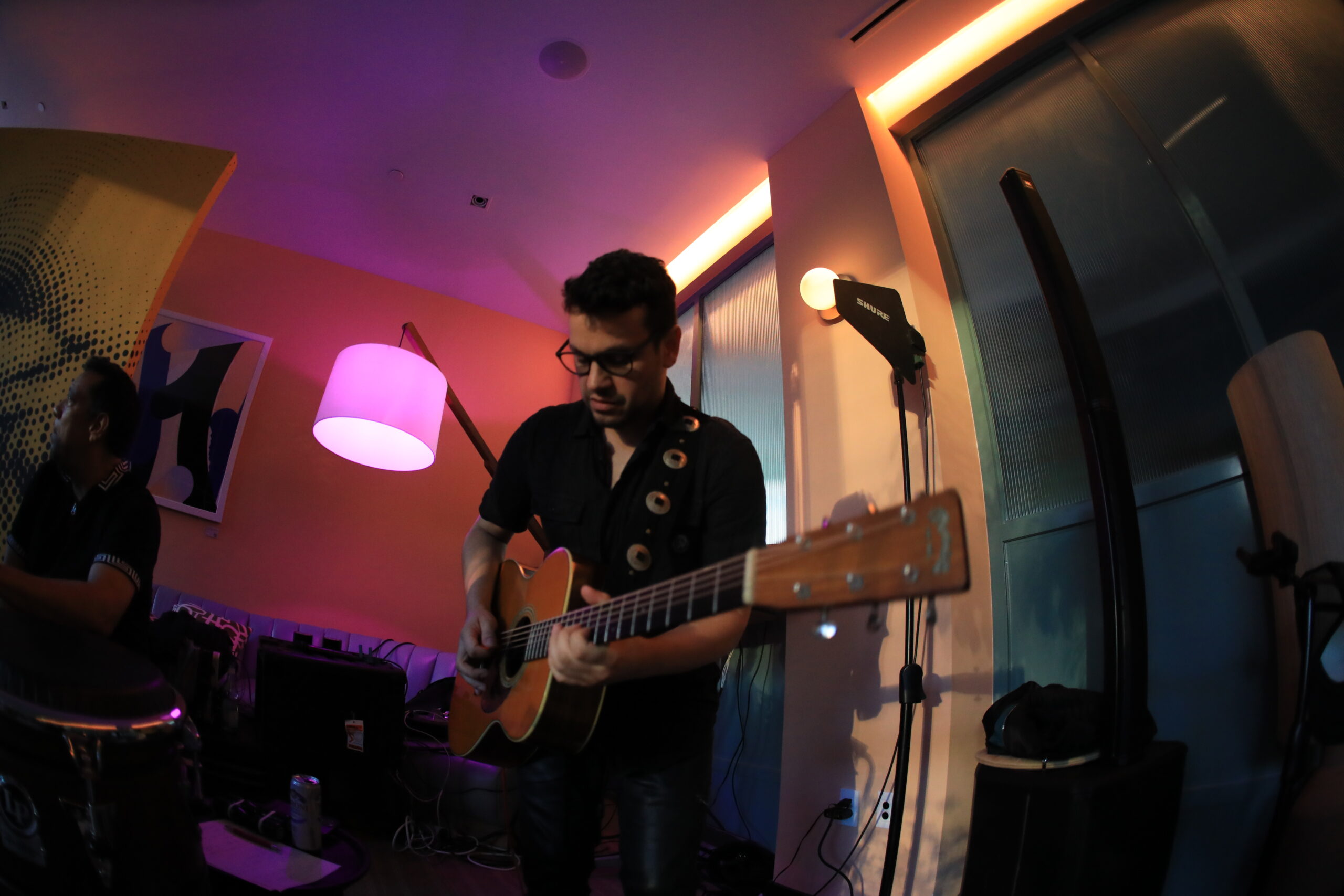 A man wearing dark glasses and clothes plays an acoustic guitar in a dimly lit room with pink and orange lighting.